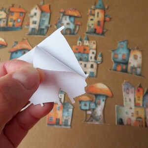 May include: A collection of whimsical house and mushroom-shaped stickers in various colors, including orange, blue, and yellow. A hand is peeling a sticker from a sheet, revealing the adhesive backing. The stickers are on a brown background.