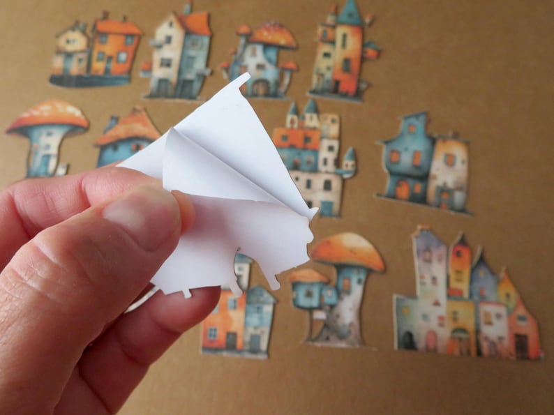 May include: A collection of whimsical house and mushroom-shaped stickers in various colors, including orange, blue, and yellow. A hand is peeling a sticker from a sheet, revealing the adhesive backing. The stickers are on a brown background.