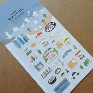 May include: A sheet of "Daily Deco Stickers" featuring travel-themed illustrations. The stickers depict landmarks, food, and transportation from Busan, South Korea, including a lighthouse, buildings, and a trolley. The color palette includes blue, white, green, and brown.