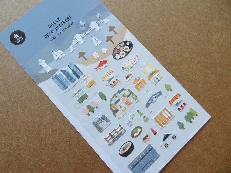 May include: A sheet of "Daily Deco Stickers" featuring travel-themed illustrations. The stickers depict landmarks, food, and transportation from Busan, South Korea, including a lighthouse, buildings, and a trolley. The color palette includes blue, white, green, and brown.