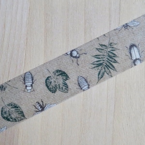 May include: Decorative washi tape featuring a repeating pattern of insects and leaves in shades of green and white on a tan background. The tape is angled across a light wood surface.