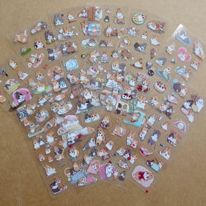 May include: Sheets of clear stickers featuring various cartoon corgi dog designs. The stickers depict corgis in different poses and settings, including sleeping, eating, and wearing accessories. The stickers are in various colors, including brown, white, and black.