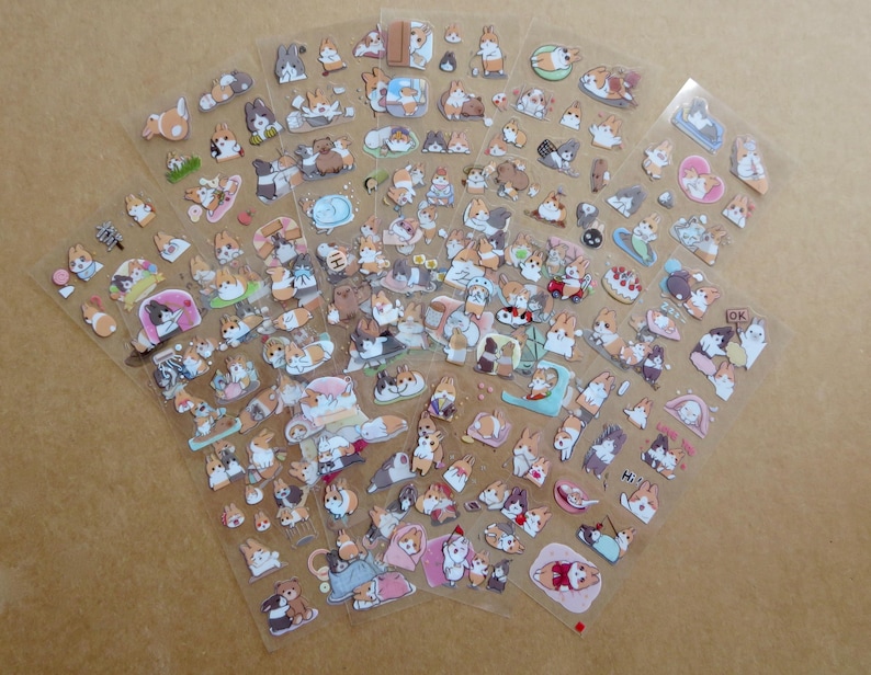 May include: Sheets of clear stickers featuring various cartoon corgi dog designs. The stickers depict corgis in different poses and settings, including sleeping, eating, and wearing accessories. The stickers are in various colors, including brown, white, and black.