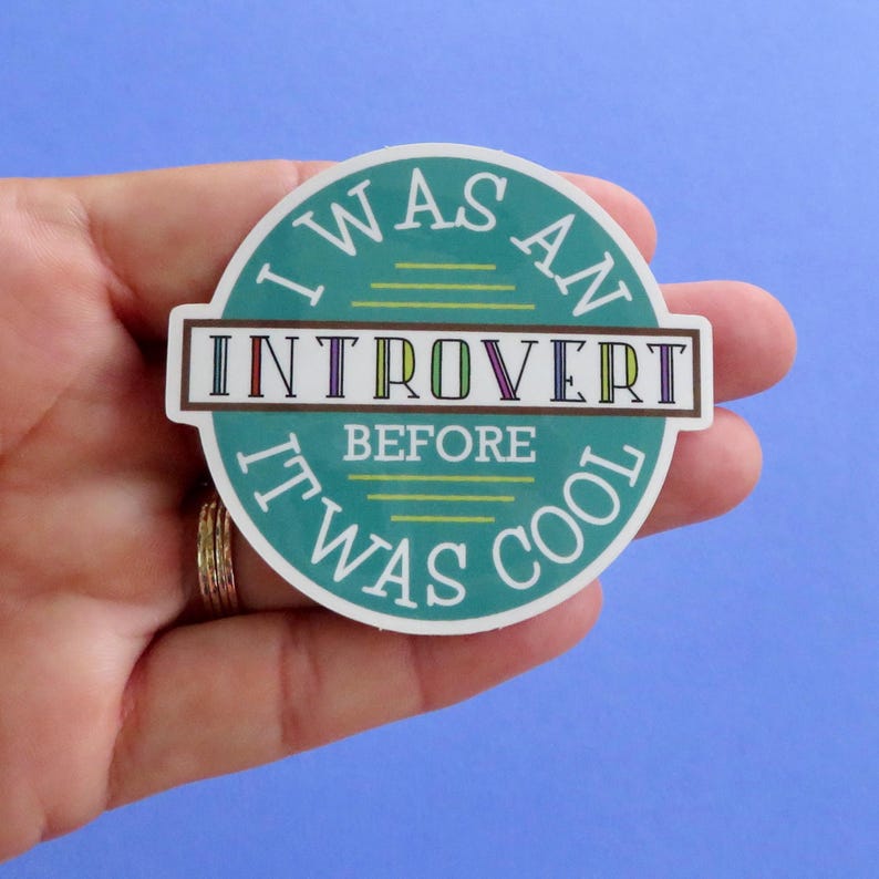 Op de afbeelding: Een ronde sticker met de tekst "I WAS AN INTROVERT BEFORE IT WAS COOL" in een turquoise en bruin ontwerp. Het woord "INTROVERT" staat in een rechthoekige doos met kleurrijke letters. De sticker wordt in een hand gehouden.