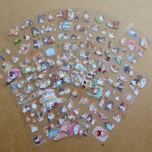 May include: Sheets of clear stickers featuring various cartoon corgi dog illustrations. The stickers depict corgis in different poses and settings, including sleeping, playing, and wearing accessories. The stickers are in shades of brown, white, and tan.