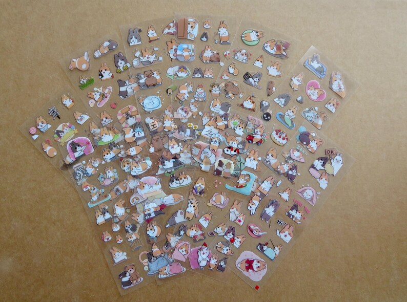 May include: Sheets of clear stickers featuring various cartoon corgi dog illustrations. The stickers depict corgis in different poses and settings, including sleeping, playing, and wearing accessories. The stickers are in shades of brown, white, and tan.