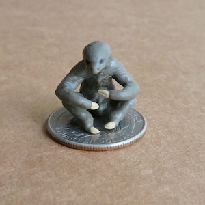 May include: A miniature grey figurine of a seated creature with crossed arms and legs. The figurine has light tan accents on its hands and feet. It is sitting on a U.S. quarter coin.