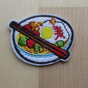 May include: Embroidered patch depicting a bowl of Asian food with chopsticks. The patch has a white border and shows a yellow egg, red and green vegetables, and a red character. The chopsticks are brown and red.