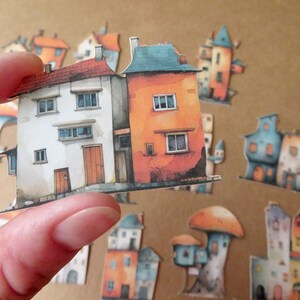 May include: A hand holding a paper cutout of two connected houses. One house is white with a red roof, the other is orange with a blue roof. Other colorful house cutouts are scattered in the background, creating a whimsical village scene.