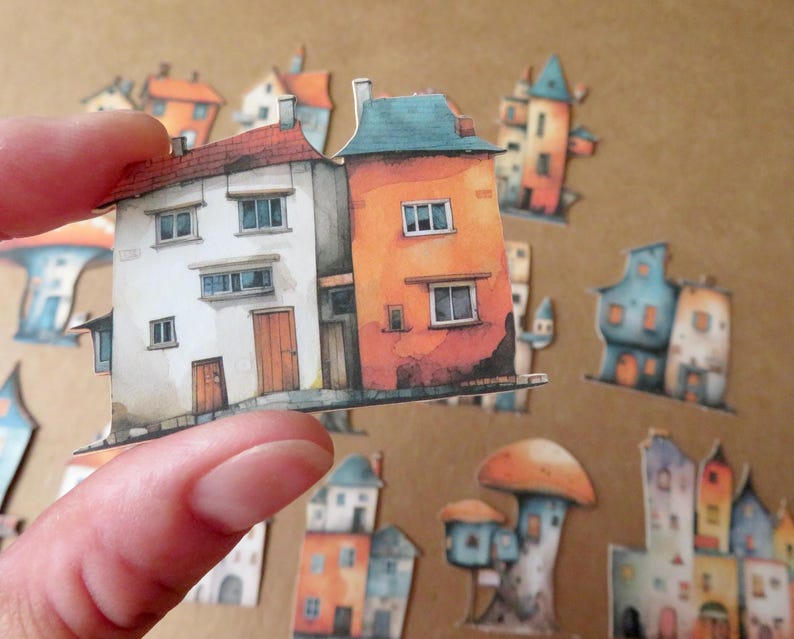 May include: A hand holding a paper cutout of two connected houses. One house is white with a red roof, the other is orange with a blue roof. Other colorful house cutouts are scattered in the background, creating a whimsical village scene.
