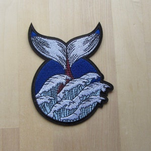 May include: Embroidered patch depicting a whale tail rising from ocean waves. The design features a blue circle, white and blue waves, and a whale tail with blue detailing. The patch has a black border.