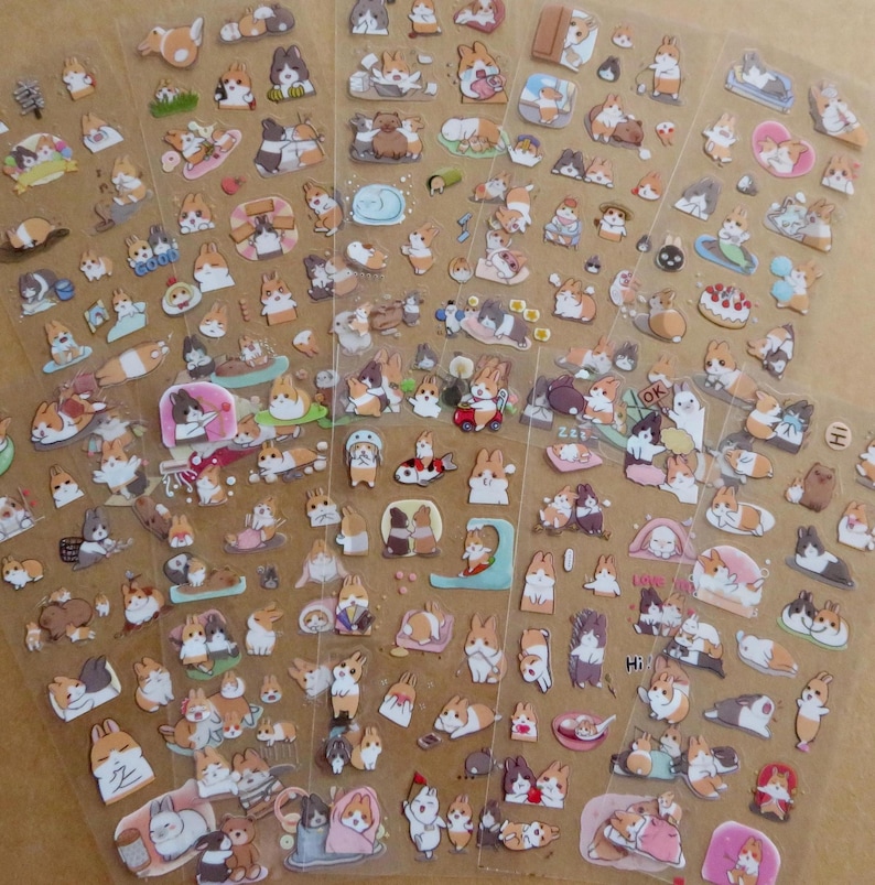 May include: Sheets of clear stickers featuring various cartoon corgi dog illustrations. The stickers depict corgis in different poses and settings, including sleeping, playing, and wearing outfits. The stickers are in shades of brown, white, and tan.