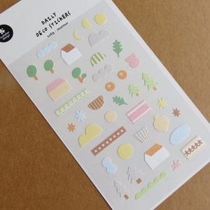 Puede incluir: Hoja de pegatinas Daily Deco con varias formas y diseños. Incluye pegatinas de casas, árboles, nubes, estrellas y otros elementos con temática de la naturaleza en colores pastel. El texto "DAILY DECO STICKERS" está impreso en la parte superior.