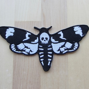 May include: A black and white embroidered patch in the shape of a moth. The moth design features a skull and skeleton-like details on the wings and body. The patch is likely for clothing or accessories.