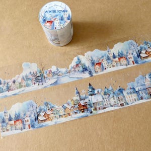 May include: A roll of decorative washi tape with a winter town scene. The tape features a watercolor illustration of snow-covered buildings, trees, and a river. The tape roll has the words "Winter Town" printed on it.