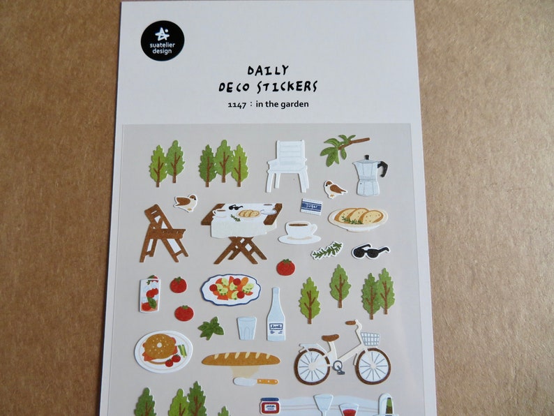 K&ouml;nnte beinhalten: Bogen mit Daily Deco-Aufklebern zum Thema Garten. Enth&auml;lt Illustrationen von B&auml;umen, St&uuml;hlen, Essen, einem Fahrrad und anderen Elementen im Freien. Der Text "Daily Deco Stickers" und "1147: in the garden" ist oben aufgedruckt.