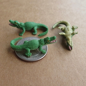 May include: Three small, green alligator figurines with open mouths and yellow eyes. One alligator is on a quarter. The alligators are approximately 2.5 cm long.