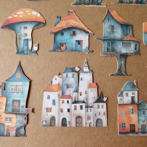 May include: A collection of whimsical paper cutouts of colorful houses and a mushroom-shaped structure. The houses feature various architectural styles with orange, blue, and white color schemes. The cutouts are arranged on a brown background.