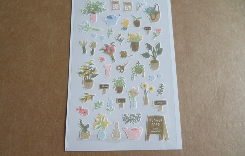 May include: A sheet of decorative stickers featuring various garden-themed illustrations. The stickers include potted plants, bouquets, gardening tools, watering cans, and signs. The stickers are in soft pastel colors, with the text "FLOWER CAFE" on one of the stickers.