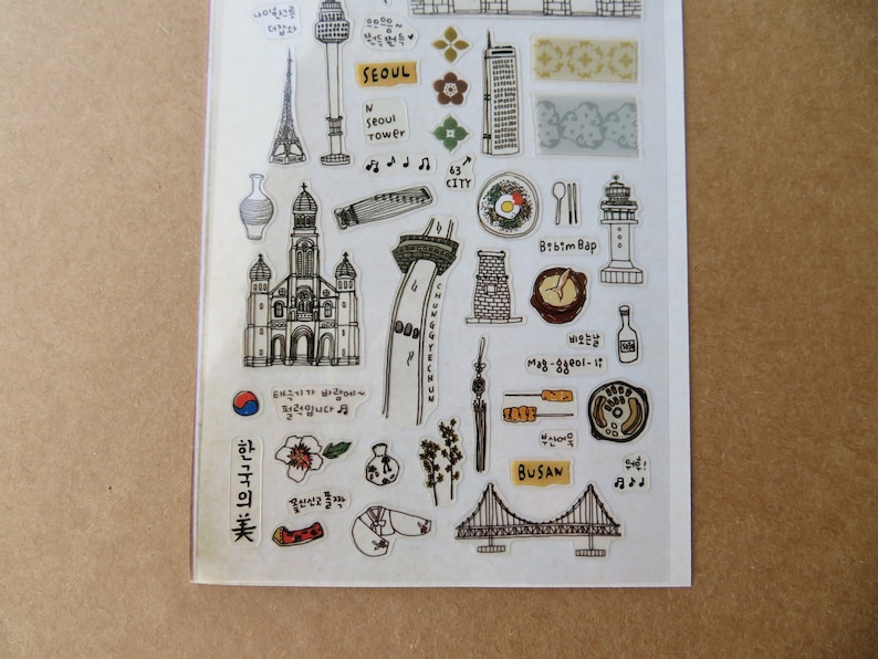May include: A clear sticker sheet featuring hand-drawn illustrations of Korean landmarks and food. Includes the Seoul Tower, a church, a bridge, and the text "SEOUL" and "BUSAN". Also features Korean text and musical notes.