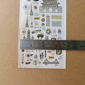May include: A clear sticker sheet featuring various black and white illustrations of Korean landmarks and cultural elements. The sheet includes images of a tower, a bridge, and food items. A ruler shows the sheet is approximately 4 inches wide.