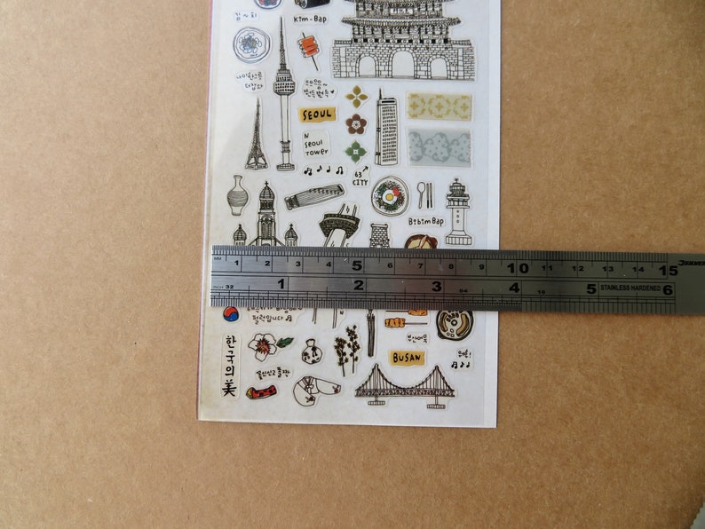 May include: A clear sticker sheet featuring various black and white illustrations of Korean landmarks and cultural elements. The sheet includes images of a tower, a bridge, and food items. A ruler shows the sheet is approximately 4 inches wide.