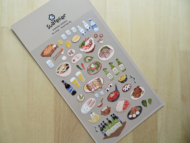 May include: A sheet of Suatelier stickers with food illustrations. The stickers depict sushi, beer, bottles, and other dishes. The text "Suatelier stickers no.1109 food trip #3" is printed on the top of the sheet.