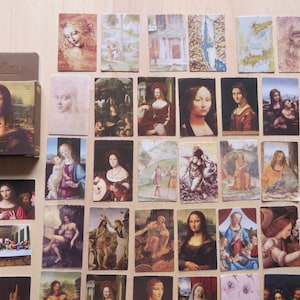 Könnte beinhalten: Eine Sammlung rechteckiger Kunstkarten mit Renaissance-Gemälden, darunter die Mona Lisa und andere Werke von Leonardo da Vinci. Eine kleine Schachtel mit dem Mona Lisa-Bild ist ebenfalls vorhanden. Die Karten sind auf einer hellbraunen Oberfläche angeordnet.