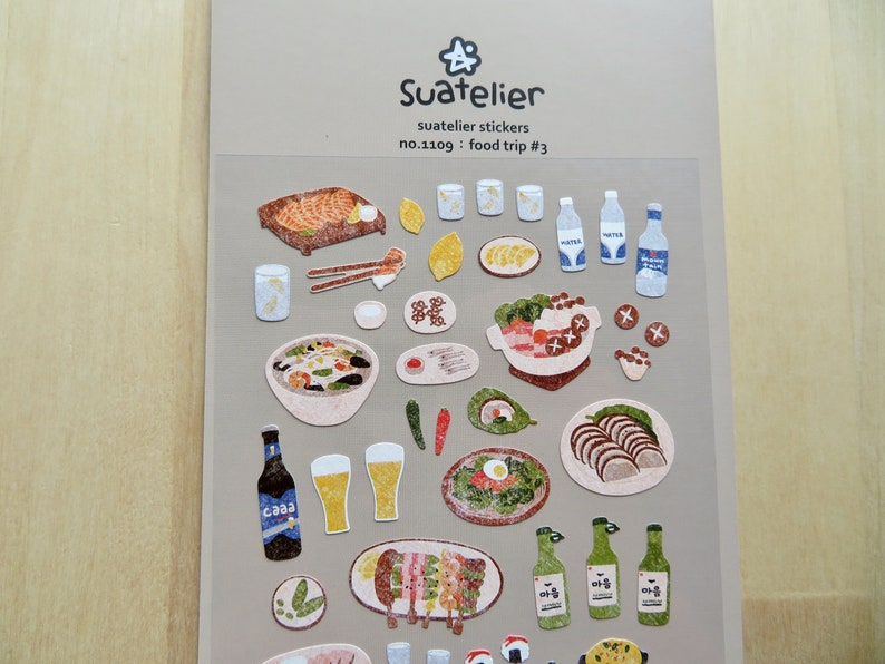May include: A sheet of Suatelier stickers with illustrations of food items such as grilled meat, bowls of soup, sushi, and bottles of water and beer. The text "Suatelier stickers" and "Food trip #3" are visible.