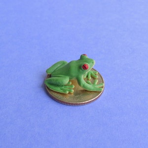 May include: A small, green frog figurine with red eyes rests on a gold-coloured coin. The frog is detailed with visible legs and a textured surface. The coin is a United States quarter.
