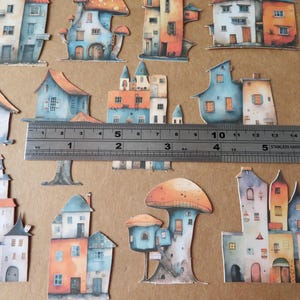 May include: Watercolor paper cutouts of whimsical houses and mushroom-shaped structures in shades of blue, orange, and white. A metal ruler shows measurements in inches. The theme is fantasy or fairy tale.