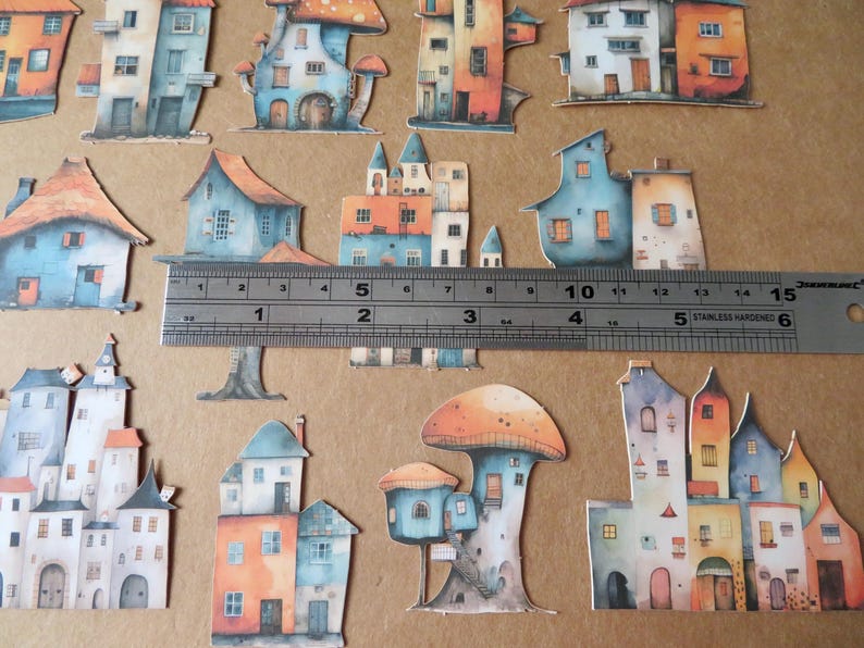 May include: Watercolor paper cutouts of whimsical houses and mushroom-shaped structures in shades of blue, orange, and white. A metal ruler shows measurements in inches. The theme is fantasy or fairy tale.