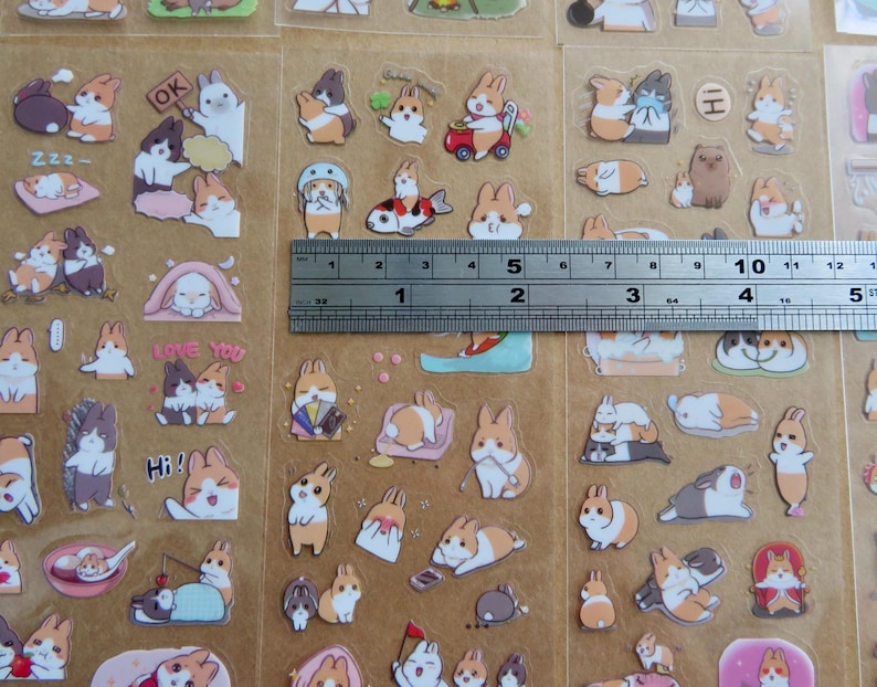 May include: Sheets of colorful bunny stickers with various poses and accessories. The stickers include bunnies with hats, fish, and text like "OK" and "Love You". A ruler shows the stickers are approximately 5 inches wide.