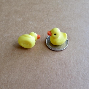 May include: Two small, yellow rubber duck figurines with orange beaks and black eyes. One duck is upright on a coin, the other is on its side. The word "CHINA" is visible on the bottom of one duck.