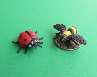 May include: A red ladybug with black spots and a black head is next to a black and yellow bee. The bee is on a quarter. Both insects are miniature figurines. The background is a solid green.