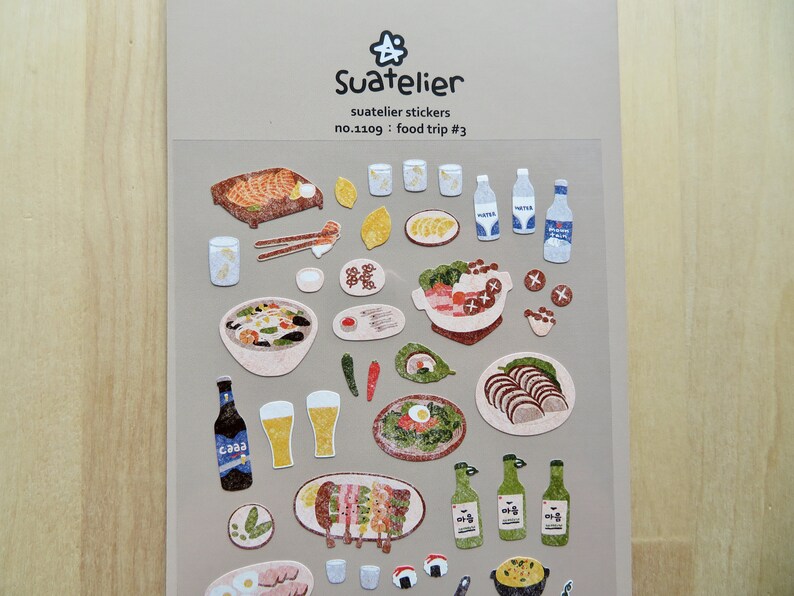 May include: A sheet of Suatelier stickers with illustrations of food items. The stickers depict dishes such as grilled fish, soup, sushi, beer, and water bottles. The text "Suatelier stickers" and "food trip #3" are displayed on the sheet.