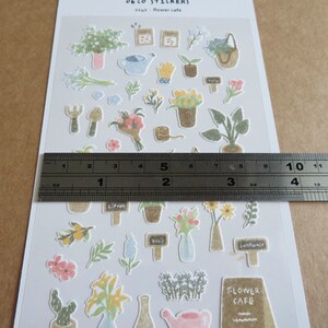 May include: A sheet of decorative stickers featuring various floral and gardening-themed illustrations. The stickers include potted plants, flowers, gardening tools, and the text "Flower Cafe". The ruler shows the sheet is approximately 5 inches long.