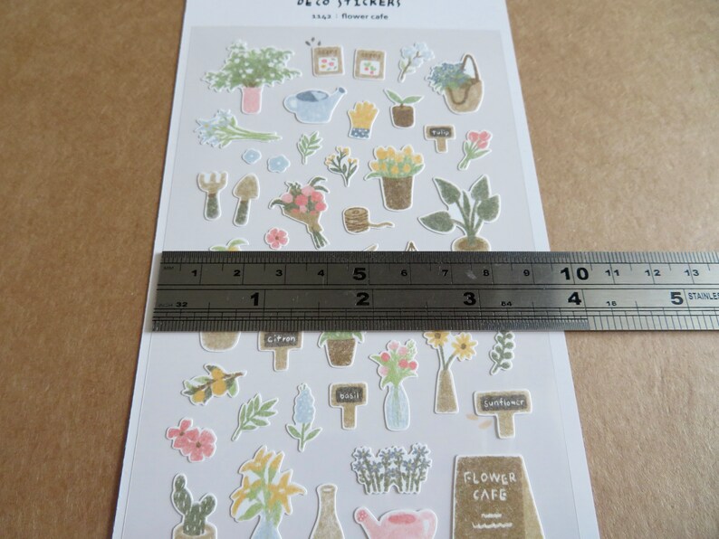 May include: A sheet of decorative stickers featuring various floral and gardening-themed illustrations. The stickers include potted plants, flowers, gardening tools, and the text "Flower Cafe". The ruler shows the sheet is approximately 5 inches long.