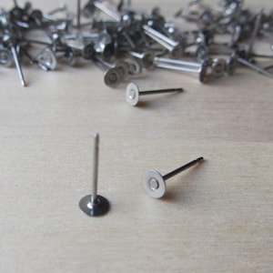 Puede incluir: Una colección de postes de pendientes plateados con almohadillas planas y circulares. Los postes están dispersos sobre una superficie de madera clara, con algunos bien enfocados. Los postes de los pendientes tienen un diseño simple y minimalista.