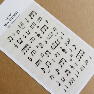 May include: A sheet of stickers featuring various black musical notes, including treble clefs, eighth notes, and quarter notes, on a light beige background with staff lines. The text "DAILY DECO STICKERS" is printed at the top.