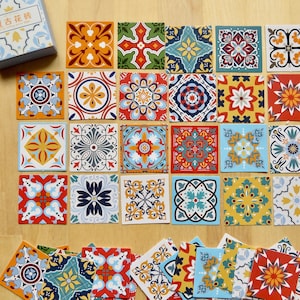 May include: A collection of colorful square tiles with intricate floral and geometric patterns. The tiles feature a variety of colors, including orange, blue, yellow, and red. A small box with Chinese text is visible in the upper left corner. The tiles are arranged on a light brown wooden surface.