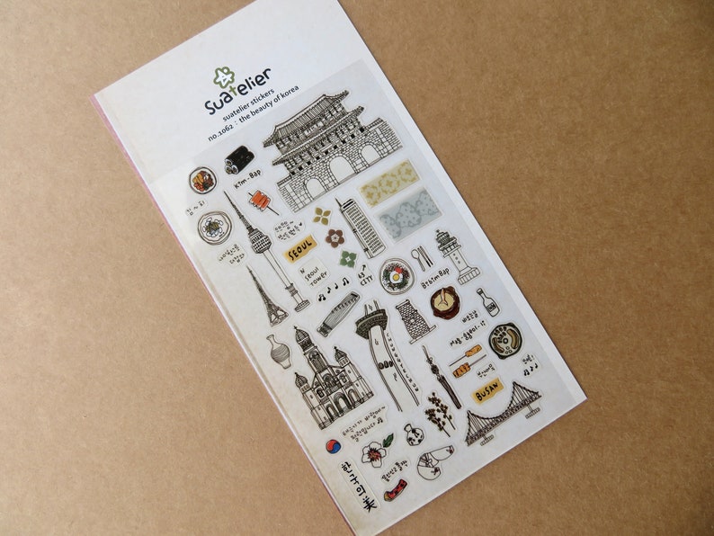 May include: A sheet of Suatelier stickers featuring black and white illustrations of Korean landmarks, food, and cultural symbols. The stickers include images of a tower, a bridge, and various food items, with text in Korean and English.