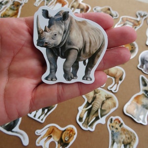 May include: A sticker of a rhinoceros with a detailed watercolour illustration. The rhino is grey with a large horn and is surrounded by other animal stickers. The stickers are on a neutral background.