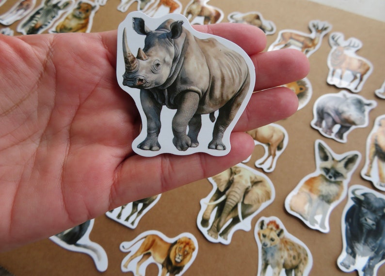 May include: A sticker of a rhinoceros with a detailed watercolour illustration. The rhino is grey with a large horn and is surrounded by other animal stickers. The stickers are on a neutral background.