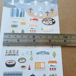 May include: Sheet of decorative stickers featuring various illustrations of buildings, food, and transportation. The stickers include images of a cityscape, a trolley, food, and a banner with Korean text. The ruler shows the sheet is approximately 6 inches long.