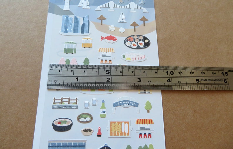 May include: Sheet of decorative stickers featuring various illustrations of buildings, food, and transportation. The stickers include images of a cityscape, a trolley, food, and a banner with Korean text. The ruler shows the sheet is approximately 6 inches long.