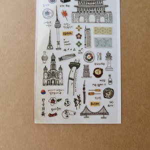 May include: A clear sticker sheet featuring black and white illustrations of Korean landmarks, food, and cultural symbols. Includes the Seoul Tower, a church, a bridge, and the word "SEOUL". The sheet has a brown background.