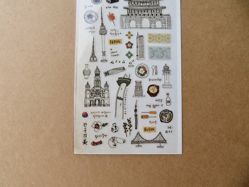 May include: A clear sticker sheet featuring black and white illustrations of Korean landmarks, food, and cultural symbols. Includes the Seoul Tower, a church, a bridge, and the word "SEOUL". The sheet has a brown background.