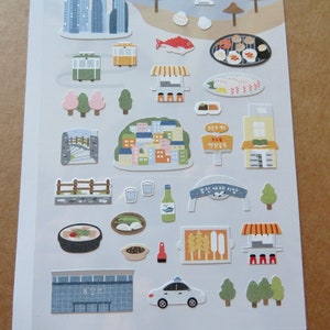 May include: A sheet of colorful stickers featuring various illustrations of buildings, food, and transportation. Includes images of a cityscape, a cable car, a taxi, and various food items. The stickers are in a cartoon style.