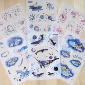 May include: A collection of sticker sheets featuring watercolor illustrations. Designs include whales, planets, moons, clouds, and sailboats in shades of blue, purple, and pink. The stickers have a dreamy, celestial theme, perfect for scrapbooking or decorating.